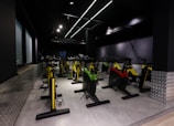 A dimly lit indoor cycling studio with multiple stationary bikes arranged in rows. The bikes have bright colors, including yellow, red, and green, contrasting with the dark walls and ceiling. The room has a modern, industrial feel with metal panels along the lower walls and sleek overhead lighting.