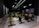 A dimly lit indoor cycling studio with multiple stationary bikes arranged in rows. The bikes have bright colors, including yellow, red, and green, contrasting with the dark walls and ceiling. The room has a modern, industrial feel with metal panels along the lower walls and sleek overhead lighting.