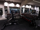 A gym with an array of dumbbells neatly organized on racks in front of large mirrors, with various strength training equipment including a bench press visible. The lighting is subtle and modern, with black and metallic gray tones dominating the space.