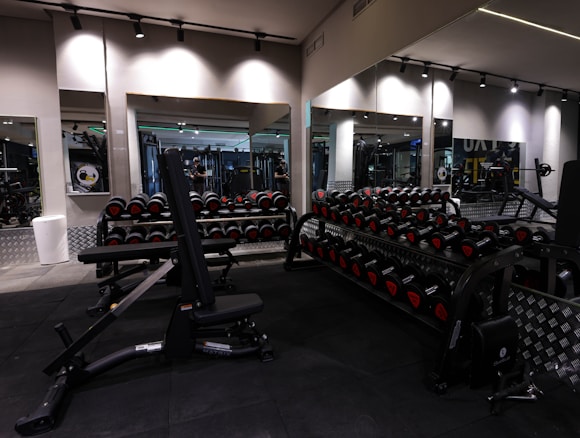 A gym with an array of dumbbells neatly organized on racks in front of large mirrors, with various strength training equipment including a bench press visible. The lighting is subtle and modern, with black and metallic gray tones dominating the space.