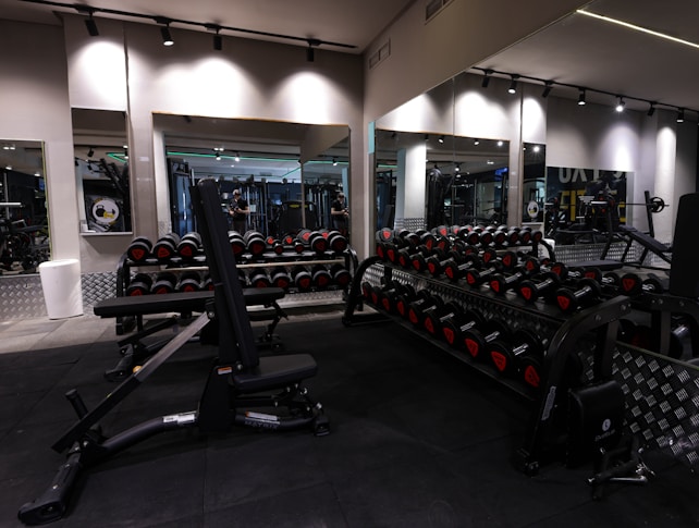 A gym with an array of dumbbells neatly organized on racks in front of large mirrors, with various strength training equipment including a bench press visible. The lighting is subtle and modern, with black and metallic gray tones dominating the space.