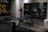 A gym setting with various workout equipment, including a weight bench with barbell and weights. The walls have a light industrial look with checkered metal paneling and motivational text partially visible. Large mirrors reflect the equipment, creating a sense of depth.