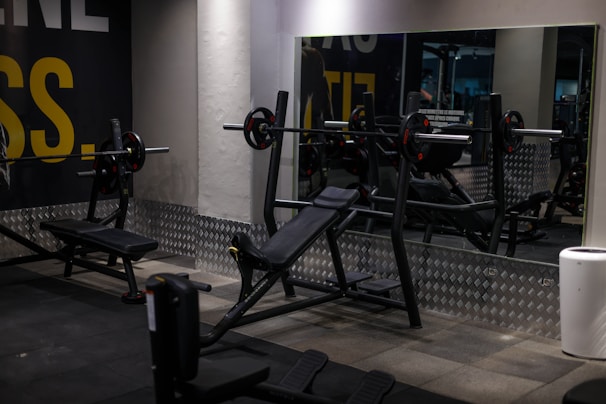 A gym setting with various workout equipment, including a weight bench with barbell and weights. The walls have a light industrial look with checkered metal paneling and motivational text partially visible. Large mirrors reflect the equipment, creating a sense of depth.