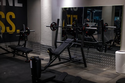 A gym setting with various workout equipment, including a weight bench with barbell and weights. The walls have a light industrial look with checkered metal paneling and motivational text partially visible. Large mirrors reflect the equipment, creating a sense of depth.