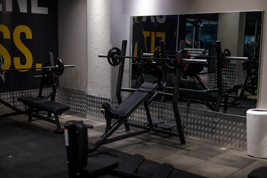 A gym setting with various workout equipment, including a weight bench with barbell and weights. The walls have a light industrial look with checkered metal paneling and motivational text partially visible. Large mirrors reflect the equipment, creating a sense of depth.