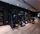 A modern gym setting filled with exercise machines such as ellipticals, stationary bikes, and weightlifting equipment. The room features a sleek design with dark walls, wooden flooring, and a mirrored wall that enhances the spacious feel. Ceiling lights provide illumination, creating an inviting atmosphere for workouts.