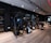 A modern gym setting filled with exercise machines such as ellipticals, stationary bikes, and weightlifting equipment. The room features a sleek design with dark walls, wooden flooring, and a mirrored wall that enhances the spacious feel. Ceiling lights provide illumination, creating an inviting atmosphere for workouts.