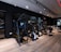A modern gym setting filled with exercise machines such as ellipticals, stationary bikes, and weightlifting equipment. The room features a sleek design with dark walls, wooden flooring, and a mirrored wall that enhances the spacious feel. Ceiling lights provide illumination, creating an inviting atmosphere for workouts.
