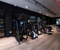A modern gym setting filled with exercise machines such as ellipticals, stationary bikes, and weightlifting equipment. The room features a sleek design with dark walls, wooden flooring, and a mirrored wall that enhances the spacious feel. Ceiling lights provide illumination, creating an inviting atmosphere for workouts.