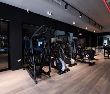 A modern gym setting filled with exercise machines such as ellipticals, stationary bikes, and weightlifting equipment. The room features a sleek design with dark walls, wooden flooring, and a mirrored wall that enhances the spacious feel. Ceiling lights provide illumination, creating an inviting atmosphere for workouts.