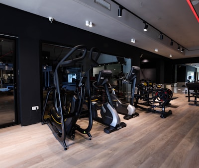 A modern gym setting filled with exercise machines such as ellipticals, stationary bikes, and weightlifting equipment. The room features a sleek design with dark walls, wooden flooring, and a mirrored wall that enhances the spacious feel. Ceiling lights provide illumination, creating an inviting atmosphere for workouts.