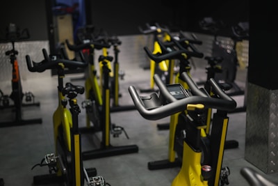 A dimly lit indoor cycling studio with multiple stationary bikes arranged in rows. The bikes are primarily yellow with black handlebars and seats. The room has a dark floor and metallic walls, and there is soft lighting creating reflections on the surfaces.