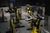 A dimly lit indoor cycling studio with multiple stationary bikes arranged in rows. The bikes are primarily yellow with black handlebars and seats. The room has a dark floor and metallic walls, and there is soft lighting creating reflections on the surfaces.