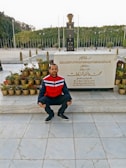 A person is crouching in front of a memorial or tombstone with inscriptions in Arabic. The area is adorned with many small potted plants, and a large structure with an eternal flame on top is seen in the background. It is an outdoor setting with a lot of greenery and trees visible in the background.