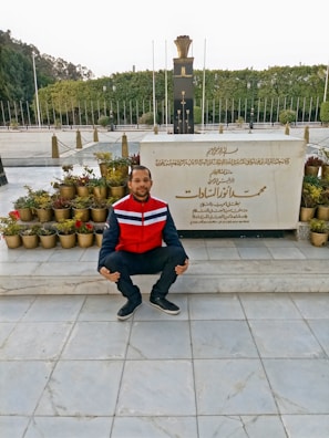 A person is crouching in front of a memorial or tombstone with inscriptions in Arabic. The area is adorned with many small potted plants, and a large structure with an eternal flame on top is seen in the background. It is an outdoor setting with a lot of greenery and trees visible in the background.