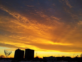 A dramatic and vivid sunset with a sky filled with golden and orange hues. Silhouettes of buildings and a unique sculpture create a striking contrast against the brightly colored sky.