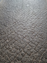 A pattern of cobblestone pavement with irregularly shaped stones in a tightly packed arrangement. The surface displays a textured and rugged appearance, with a mix of gray and earthy tones.