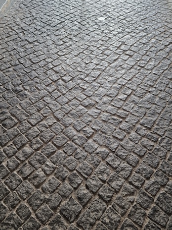 A pattern of cobblestone pavement with irregularly shaped stones in a tightly packed arrangement. The surface displays a textured and rugged appearance, with a mix of gray and earthy tones.