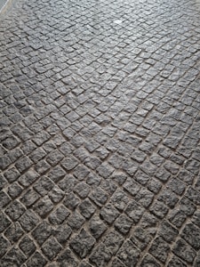A pattern of cobblestone pavement with irregularly shaped stones in a tightly packed arrangement. The surface displays a textured and rugged appearance, with a mix of gray and earthy tones.