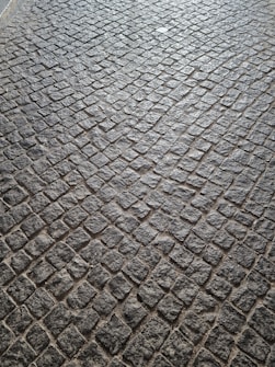 A pattern of cobblestone pavement with irregularly shaped stones in a tightly packed arrangement. The surface displays a textured and rugged appearance, with a mix of gray and earthy tones.