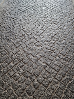 A pattern of cobblestone pavement with irregularly shaped stones in a tightly packed arrangement. The surface displays a textured and rugged appearance, with a mix of gray and earthy tones.