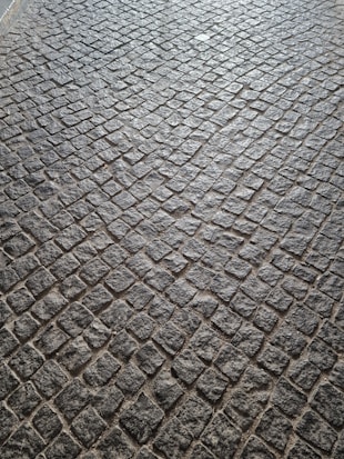 A pattern of cobblestone pavement with irregularly shaped stones in a tightly packed arrangement. The surface displays a textured and rugged appearance, with a mix of gray and earthy tones.