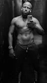 A muscular man stands shirtless in a narrow, dimly lit hallway. He holds a smartphone in one hand, capturing a mirror selfie. He wears athletic pants and a watch on his wrist. The environment appears industrial with a dark ambiance.