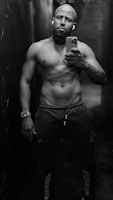 A muscular man stands shirtless in a narrow, dimly lit hallway. He holds a smartphone in one hand, capturing a mirror selfie. He wears athletic pants and a watch on his wrist. The environment appears industrial with a dark ambiance.