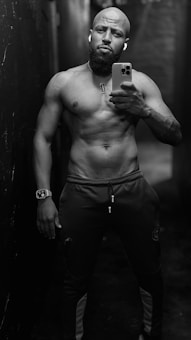 A muscular man stands shirtless in a narrow, dimly lit hallway. He holds a smartphone in one hand, capturing a mirror selfie. He wears athletic pants and a watch on his wrist. The environment appears industrial with a dark ambiance.