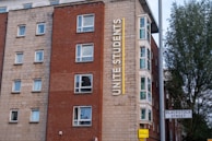 A multi-story building with a red and beige brick facade features multiple square windows. The building has a large vertical sign that reads 'Unite Students' in yellow letters. A street sign indicates 'Blackstock Street' in front of the building, and there is a tree visible on the right side.