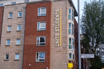 A multi-story building with a red and beige brick facade features multiple square windows. The building has a large vertical sign that reads 'Unite Students' in yellow letters. A street sign indicates 'Blackstock Street' in front of the building, and there is a tree visible on the right side.