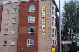 A multi-story building with a red and beige brick facade features multiple square windows. The building has a large vertical sign that reads 'Unite Students' in yellow letters. A street sign indicates 'Blackstock Street' in front of the building, and there is a tree visible on the right side.
