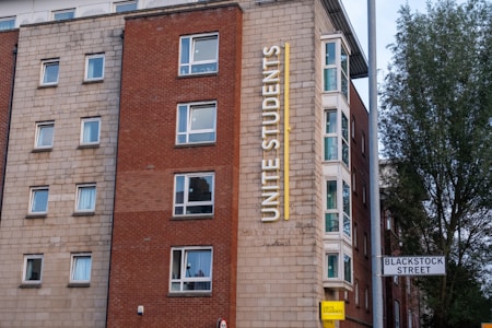 A multi-story building with a red and beige brick facade features multiple square windows. The building has a large vertical sign that reads 'Unite Students' in yellow letters. A street sign indicates 'Blackstock Street' in front of the building, and there is a tree visible on the right side.