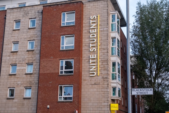 A multi-story building with a red and beige brick facade features multiple square windows. The building has a large vertical sign that reads 'Unite Students' in yellow letters. A street sign indicates 'Blackstock Street' in front of the building, and there is a tree visible on the right side.