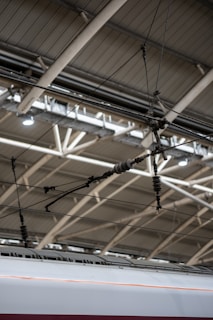 A section of a modern train station ceiling with metal beams and overhead electrical wires, above an aerodynamic high-speed train.