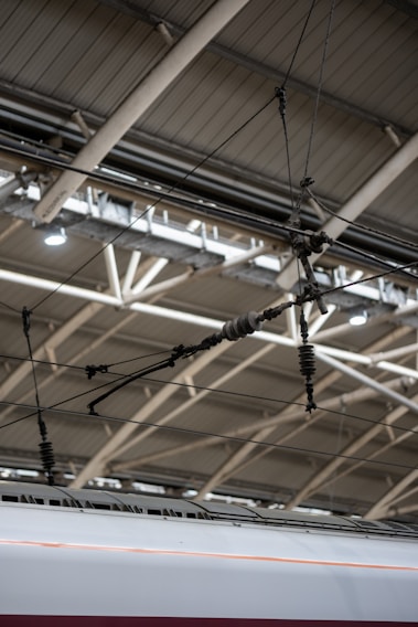 A section of a modern train station ceiling with metal beams and overhead electrical wires, above an aerodynamic high-speed train.