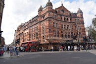 A large, ornate red brick building with intricate architectural details, likely a theater or historical building. A double-decker bus is passing by on the street, which is busy with pedestrians. The entrance features a sign related to a popular play or production.