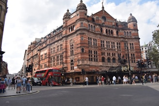 A large, ornate red brick building with intricate architectural details, likely a theater or historical building. A double-decker bus is passing by on the street, which is busy with pedestrians. The entrance features a sign related to a popular play or production.