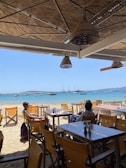 A beachside restaurant with wooden deck chairs and tables. There are two people sitting at a table, enjoying the view of the sea. The ocean is calm with several sailboats anchored in the distance. The restaurant is shaded with a woven thatch roof and straw lampshades. The sandy beach leads directly to the bright blue water.