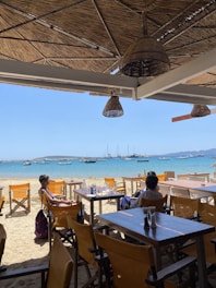 A beachside restaurant with wooden deck chairs and tables. There are two people sitting at a table, enjoying the view of the sea. The ocean is calm with several sailboats anchored in the distance. The restaurant is shaded with a woven thatch roof and straw lampshades. The sandy beach leads directly to the bright blue water.