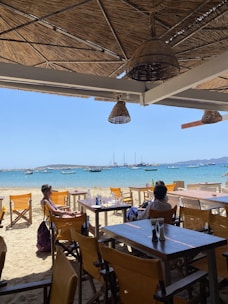 A beachside restaurant with wooden deck chairs and tables. There are two people sitting at a table, enjoying the view of the sea. The ocean is calm with several sailboats anchored in the distance. The restaurant is shaded with a woven thatch roof and straw lampshades. The sandy beach leads directly to the bright blue water.