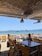 A beachside restaurant with wooden deck chairs and tables. There are two people sitting at a table, enjoying the view of the sea. The ocean is calm with several sailboats anchored in the distance. The restaurant is shaded with a woven thatch roof and straw lampshades. The sandy beach leads directly to the bright blue water.