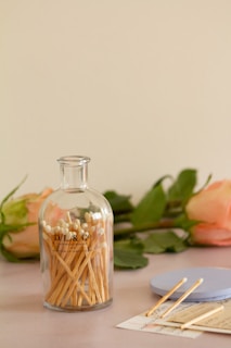 A glass bottle filled with wooden safety matches is placed on a surface. In the background, there are two peach-colored roses with green stems and leaves. Beside the bottle, there is a round blue lid and several matches lying on top of a handwritten note.