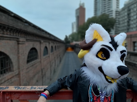 A person wearing a colorful animal costume stands in an urban environment. The costume features a large white, yellow, and black animal head with expressive eyes and fur details. The person is leaning casually on a metal railing, with their arm resting on it. The background includes brick buildings and some modern skyscrapers, suggesting a city setting.