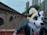 A person wearing a colorful animal costume stands in an urban environment. The costume features a large white, yellow, and black animal head with expressive eyes and fur details. The person is leaning casually on a metal railing, with their arm resting on it. The background includes brick buildings and some modern skyscrapers, suggesting a city setting.