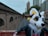 A person wearing a colorful animal costume stands in an urban environment. The costume features a large white, yellow, and black animal head with expressive eyes and fur details. The person is leaning casually on a metal railing, with their arm resting on it. The background includes brick buildings and some modern skyscrapers, suggesting a city setting.