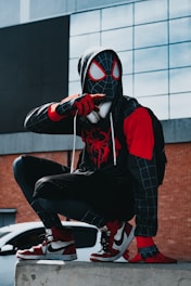 A person is dressed in a black and red superhero costume with a web pattern, wearing a mask. They are crouched on a concrete ledge in an urban setting, with a large building featuring reflective panels behind them. The person is wearing red and white sneakers, and headphones are visible around their neck.