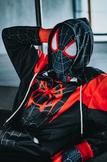 A person is wearing a black and red superhero costume with a hood and mask that features a web-like pattern and large white eye shapes. The costume includes red accents and a spider symbol on the chest. The individual is reclining with one hand resting behind their head.