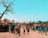 A large group of people, including many children, gather in an outdoor setting. A significant number of them are looking up at a flying drone in the sky. There are various makeshift and traditional-style structures with thatched roofs in the background. A large leafless tree is prominent on the left side of the image. The day appears sunny, enhancing the natural colors of the scene.