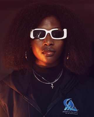 A person with curly hair is wearing oversized white sunglasses and a dark jacket with a logo. The lighting casts a warm glow on the face, highlighting glossy lips and hoop earrings. A cross necklace is visible, adding to the stylish appearance.